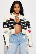Woman wearing a white jacket with logos, black bra, and blue jeans on a light gray background