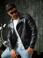 Man wearing a black leather jacket and sunglasses, standing next to a motorcycle.