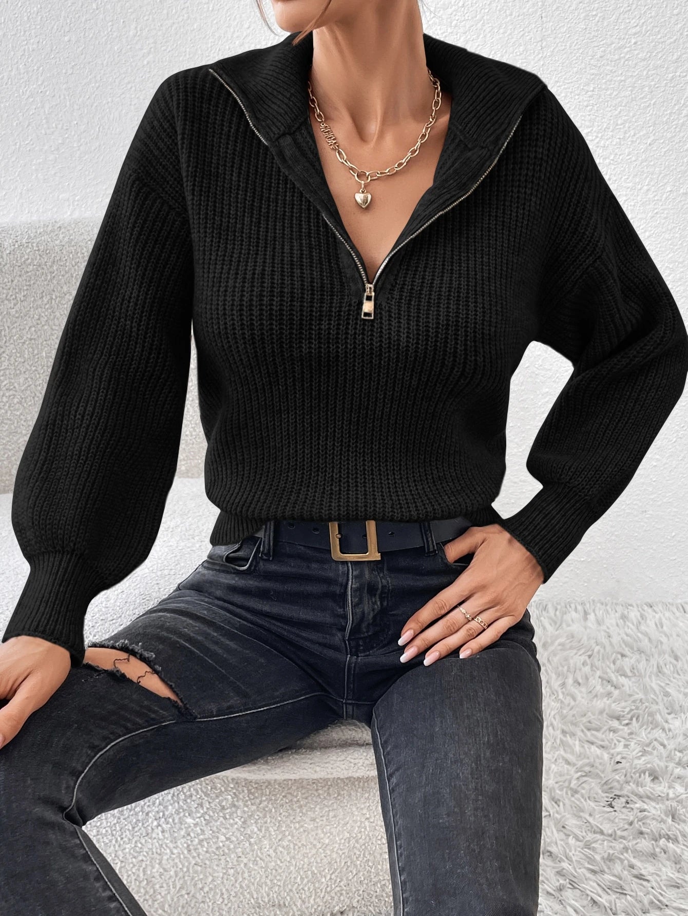 Person wearing a black ribbed sweater and dark jeans sitting on a light-colored surface.