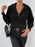 Person wearing a black zip-up sweater and black jeans, holding a black handbag against a light gray background.