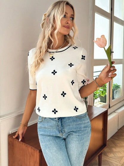 Woman wearing a white top with black patterns and blue jeans, holding a pink tulip indoors.