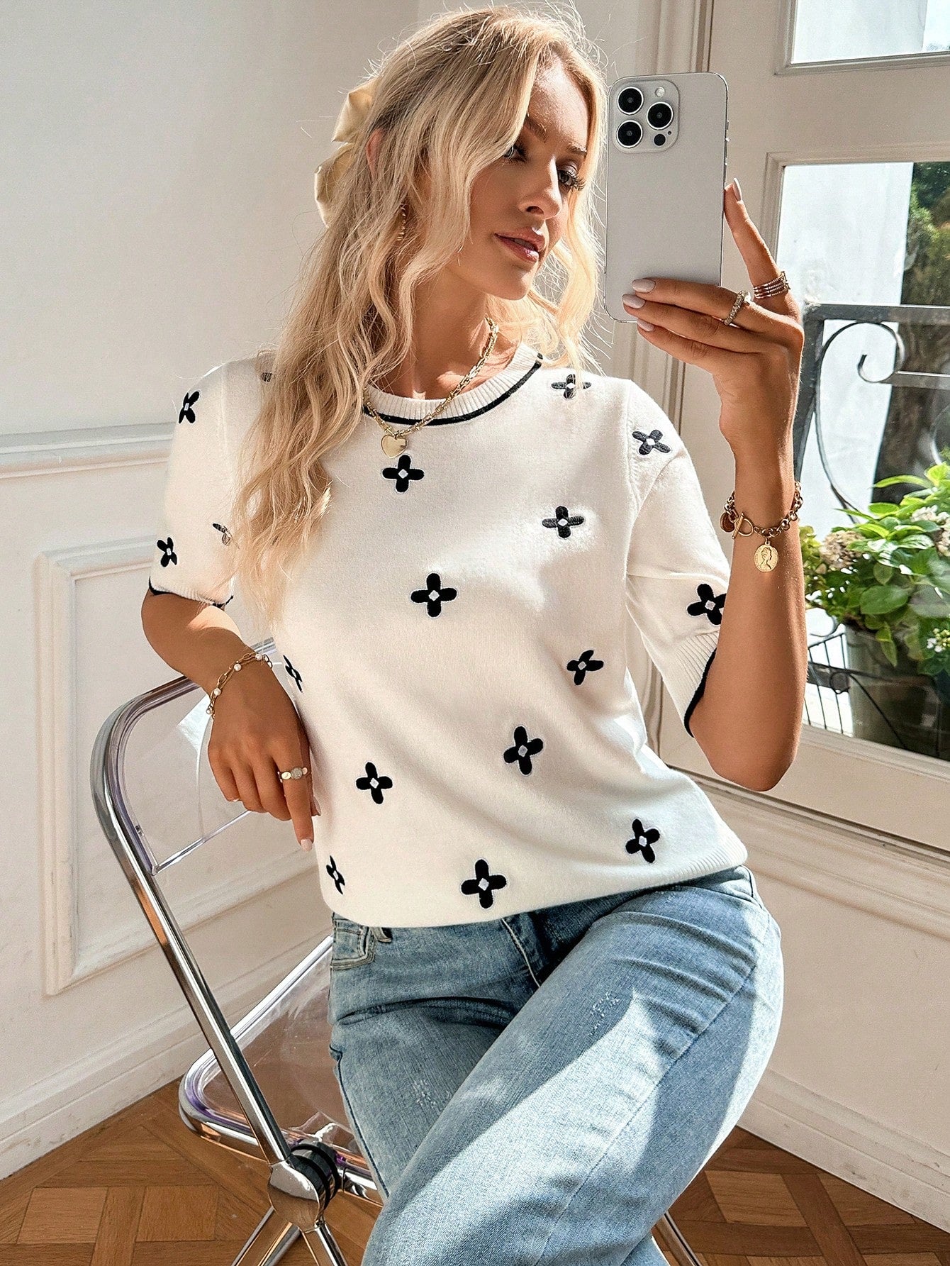 Woman taking a selfie wearing a white top with black patterns and blue jeans indoors.