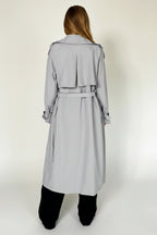 Person wearing a light gray trench coat in a minimal setting with a chair.