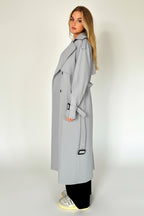 Woman wearing a light gray trench coat standing against a plain background.