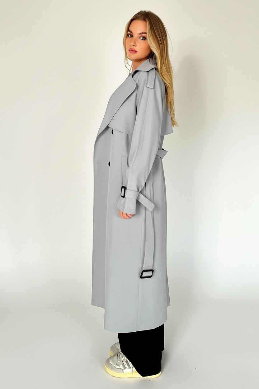 Woman wearing a light gray trench coat standing against a plain background.