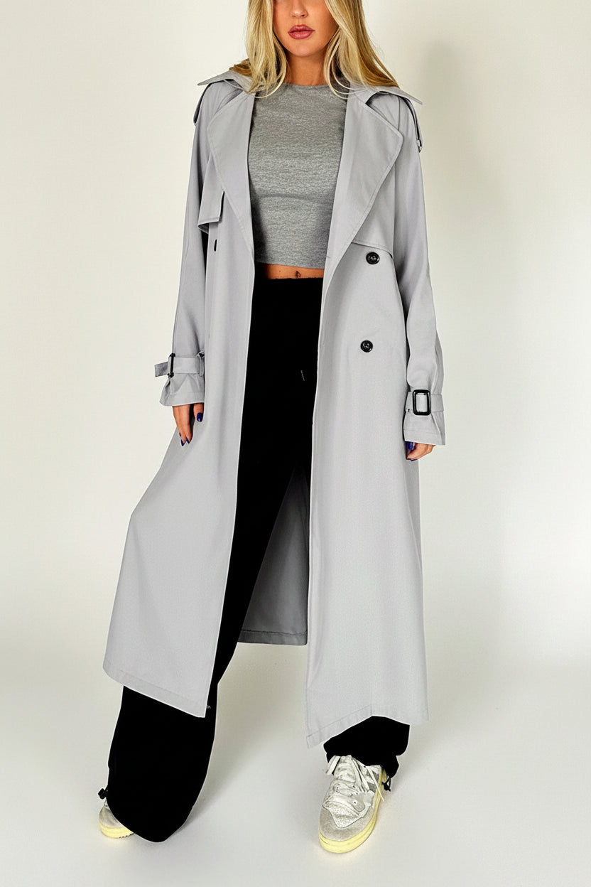 Person wearing a long gray trench coat over a gray top and black pants, standing against a plain background.