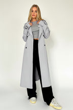 Woman wearing a light gray trench coat over a gray outfit in a minimalistic setting.