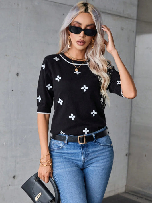 Woman wearing a black sweater with white patterns and blue jeans against a gray wall.