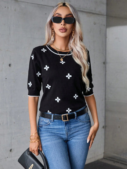 Woman wearing a black top with white patterns and blue jeans against a gray concrete wall.