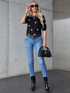 Woman wearing a black top with white patterns, blue jeans, and black boots against a concrete wall.