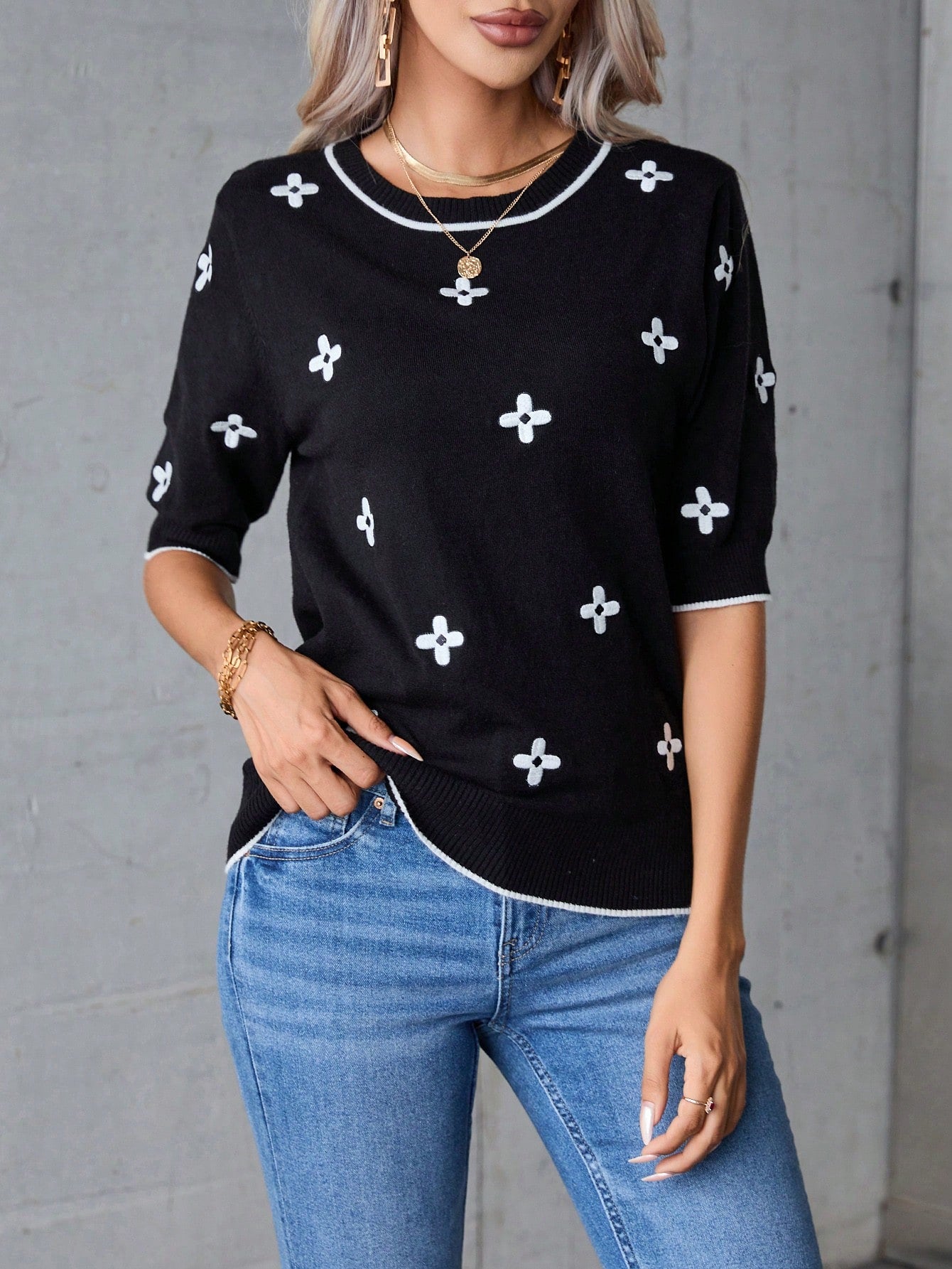 Person wearing a black top with white patterns and blue jeans against a gray background