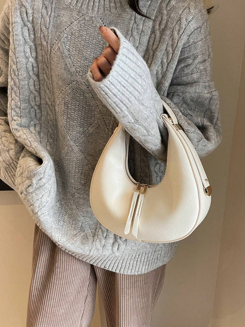 Person wearing a gray cable knit sweater holding a cream handbag.