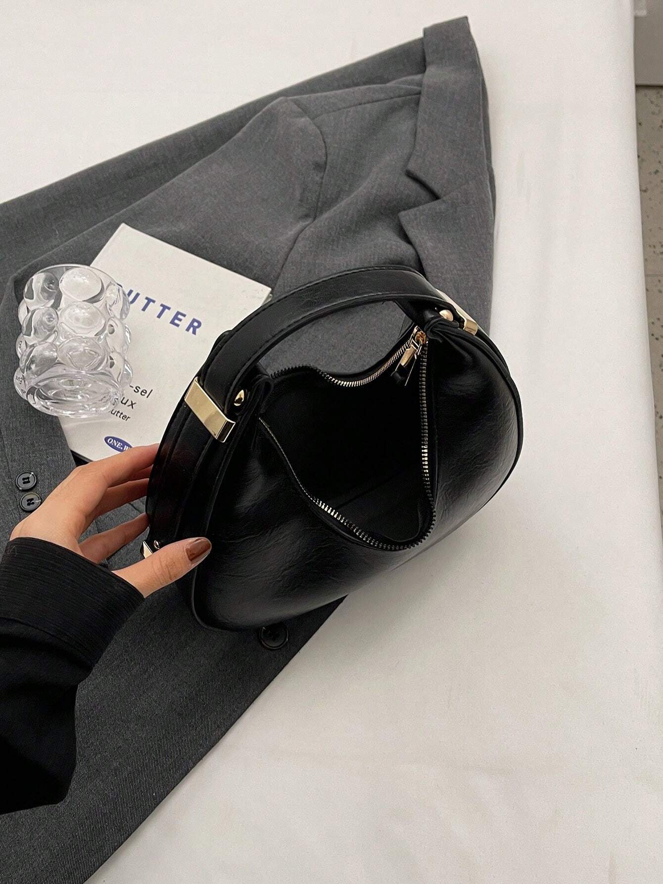 Black handbag on a white surface with a gray jacket and a glass bottle.