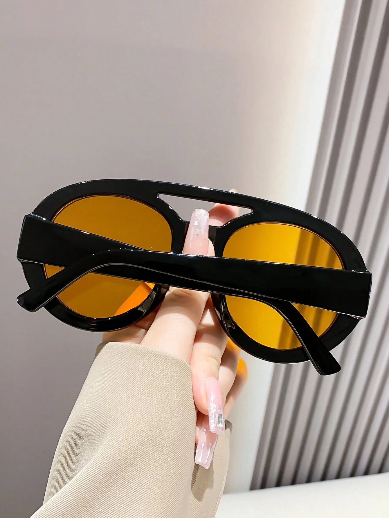 Person holding a pair of black sunglasses with yellow lenses against a neutral background