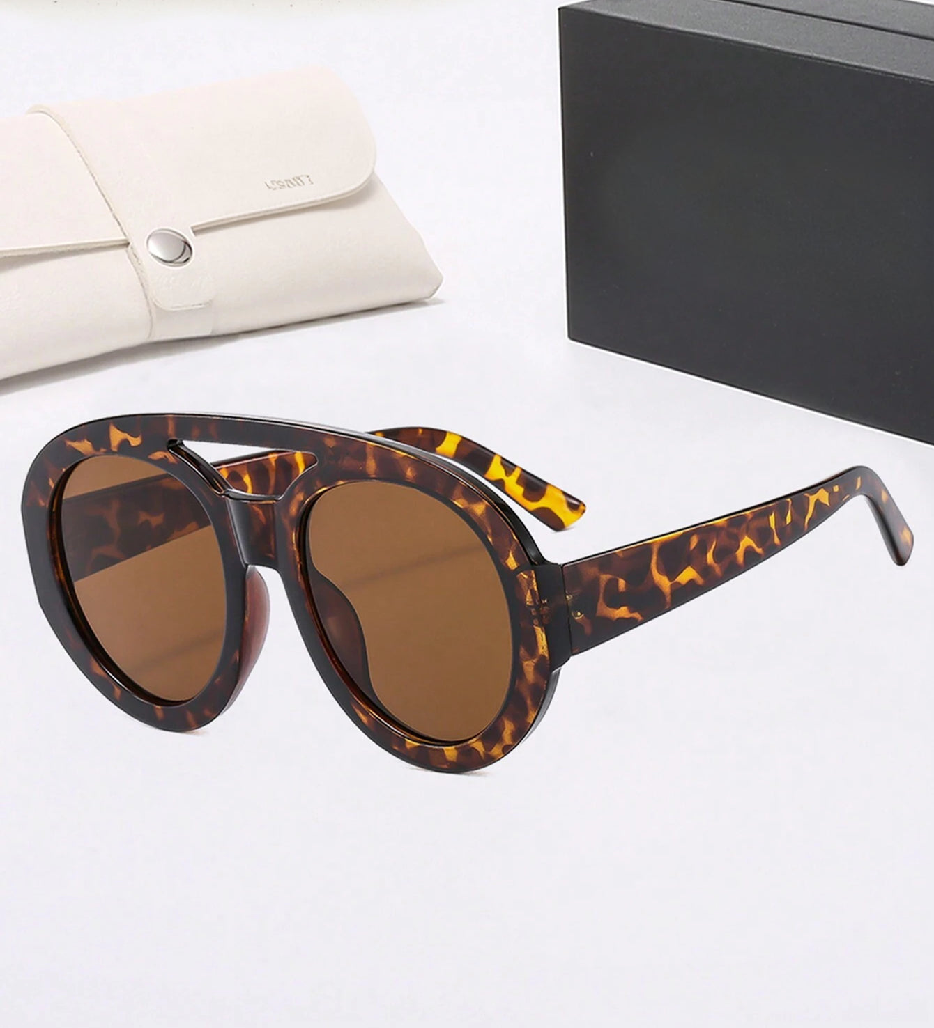 Tortoiseshell sunglasses with brown lenses on a white surface, next to a beige handbag and black box.