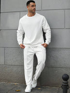 Man wearing a white tracksuit standing against a gray wall.