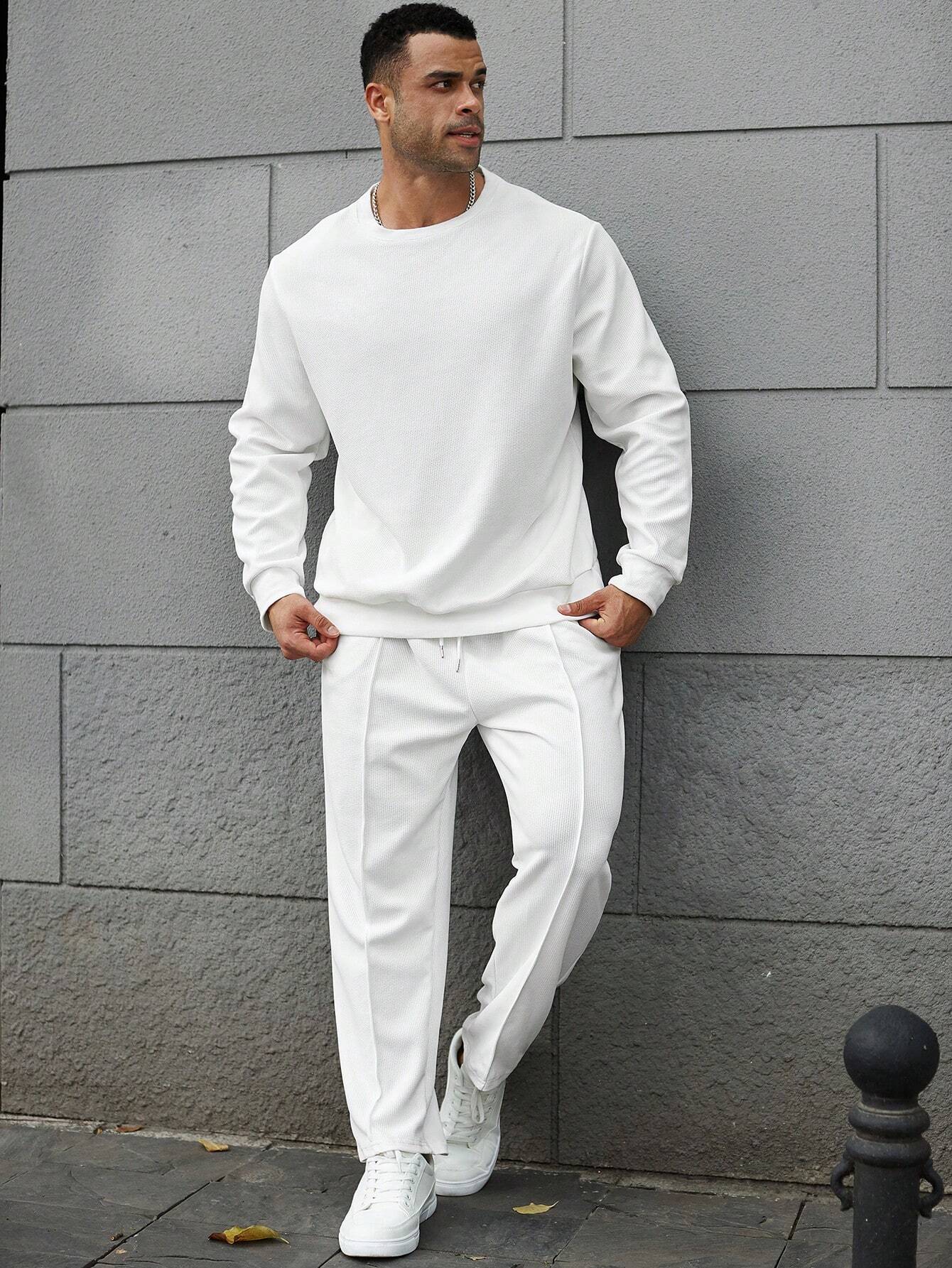 Man wearing a white tracksuit standing against a gray wall.