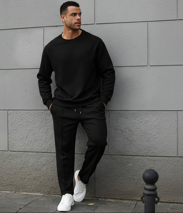 Man wearing a black tracksuit standing against a gray wall.