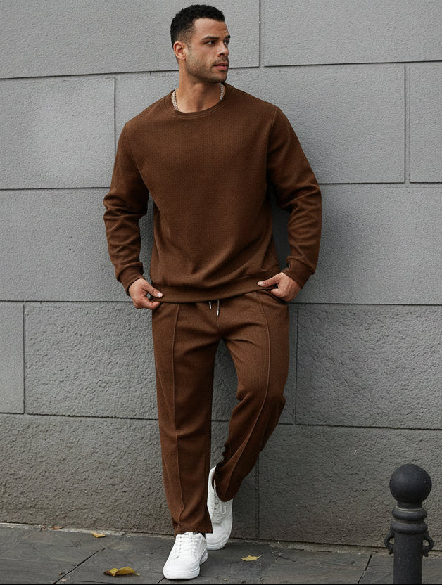 Man wearing a brown tracksuit standing against a gray wall.