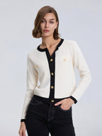 Woman wearing a white cardigan with black trim and gold buttons on a light gray background