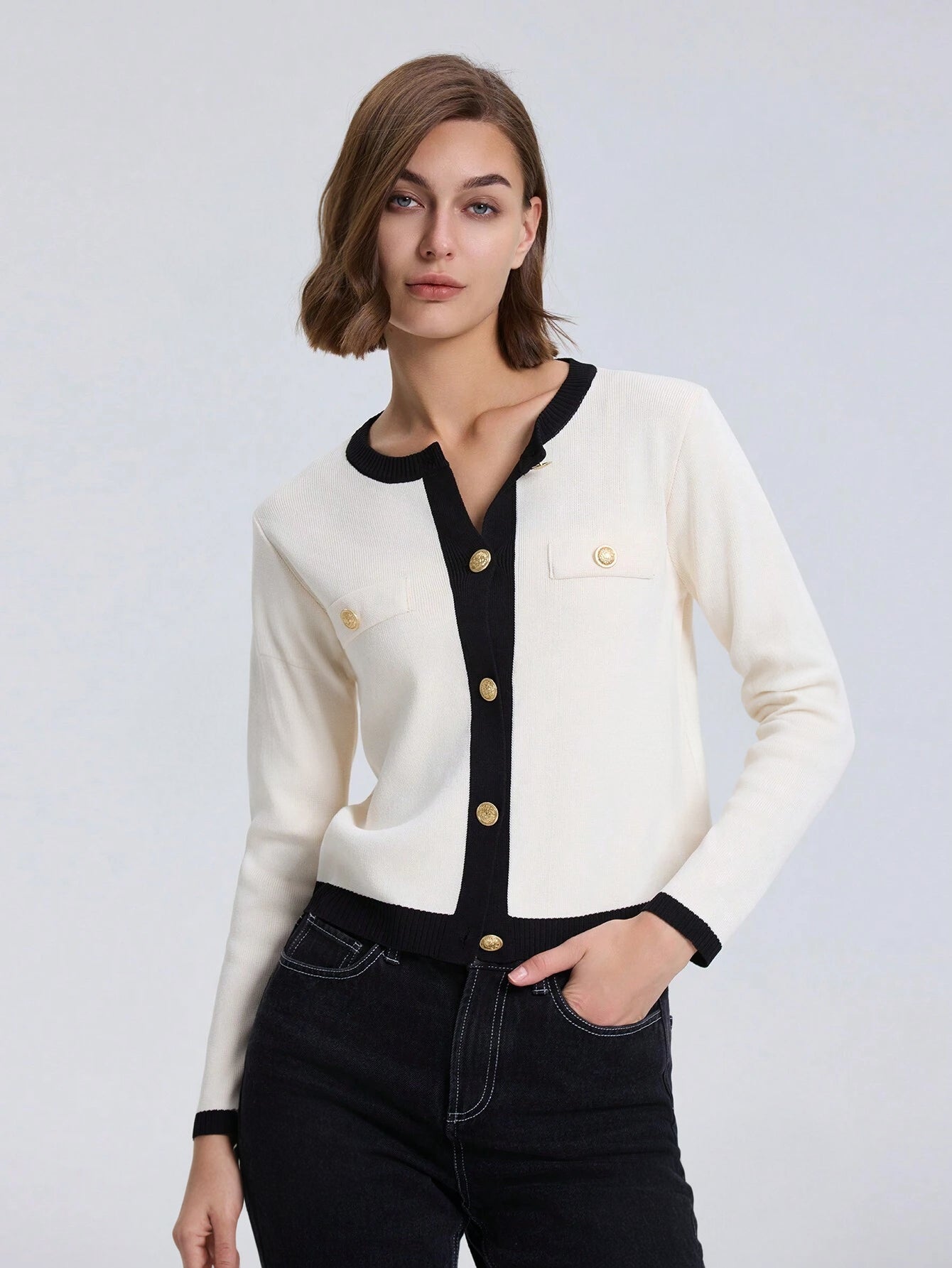 Woman wearing a white cardigan with black trim and gold buttons on a light gray background