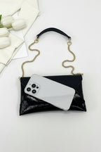 Black clutch with gold chain holding a phone on a white surface with flowers.