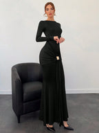 Woman wearing a black dress standing next to a black chair against a white wall.