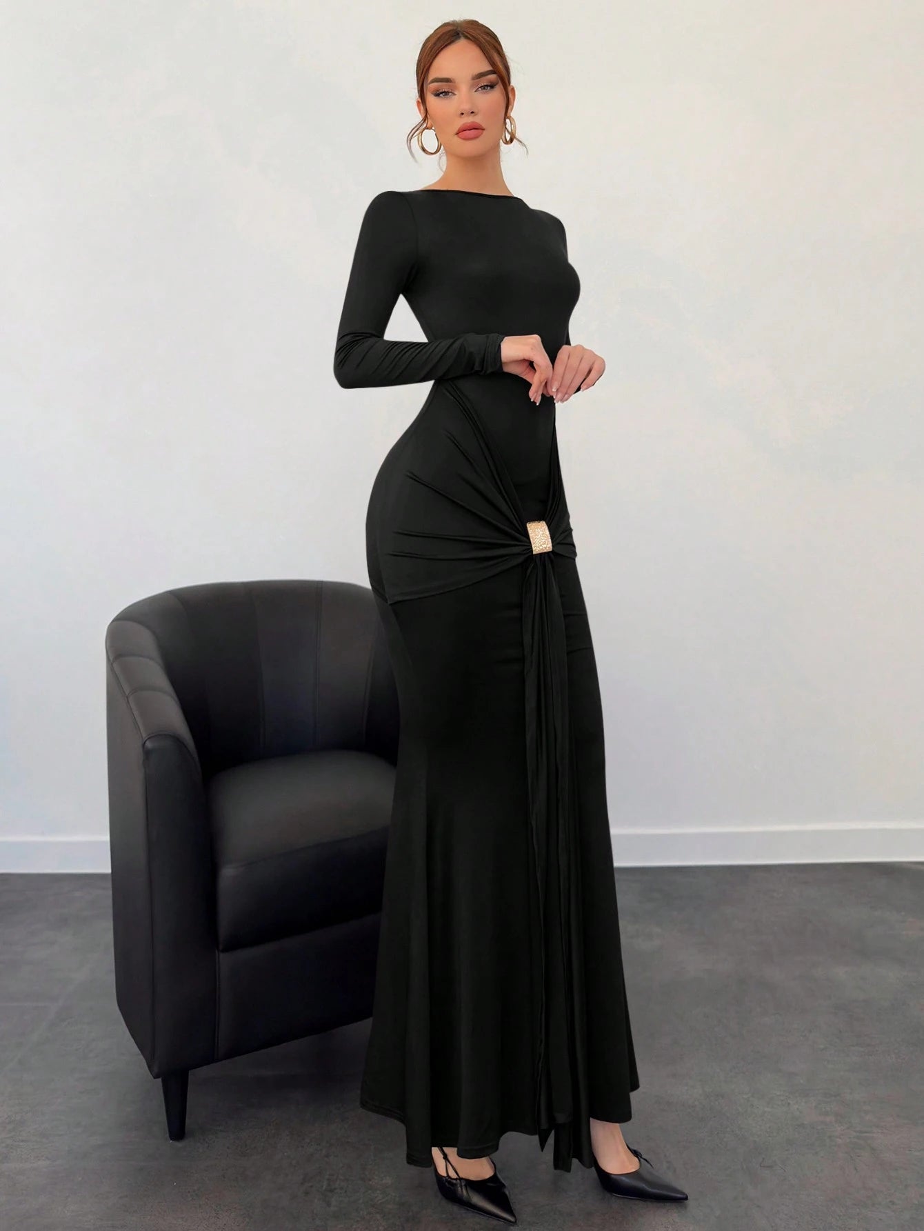 Woman wearing a black dress standing next to a black chair against a white wall.