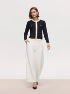 Woman wearing a black cardigan with gold buttons and white pants on a plain background