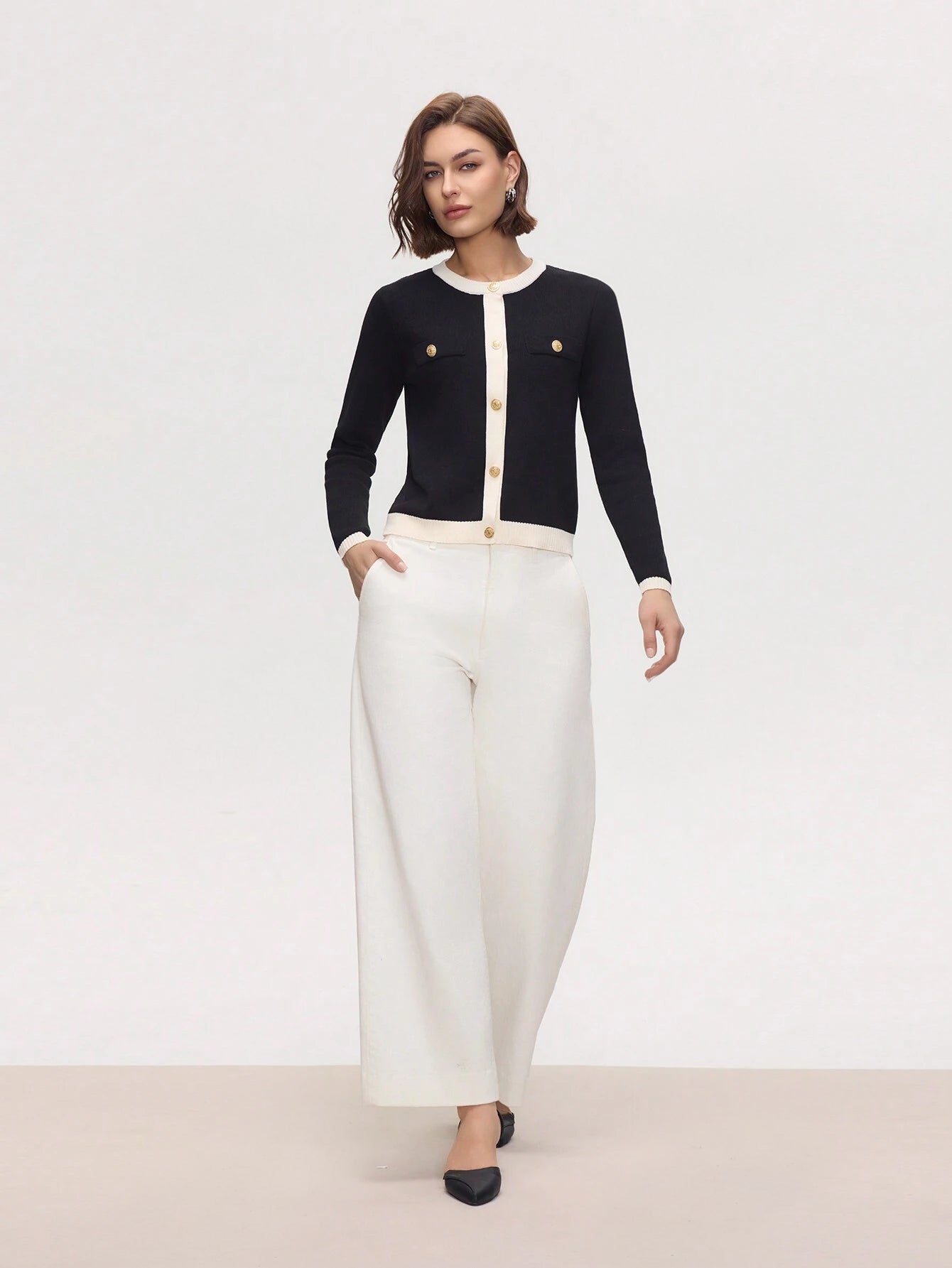 Woman wearing a black cardigan with gold buttons and white pants on a plain background
