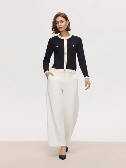 Woman wearing a black cardigan with gold buttons and white pants on a plain background