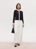Woman wearing a black cardigan, white blouse, and white pants with a black handbag on a light gray background