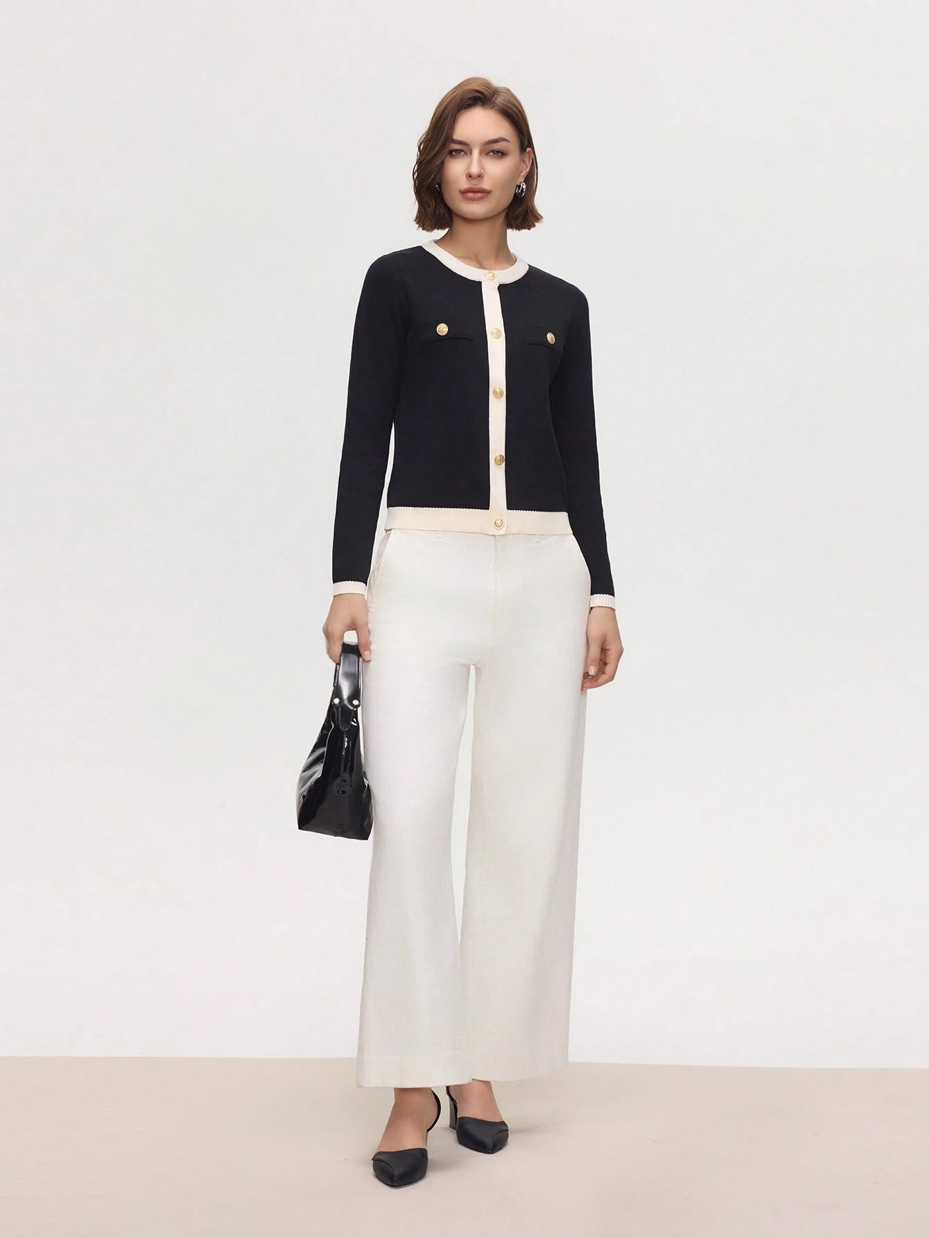 Woman wearing a black cardigan, white blouse, and white pants with a black handbag on a light gray background