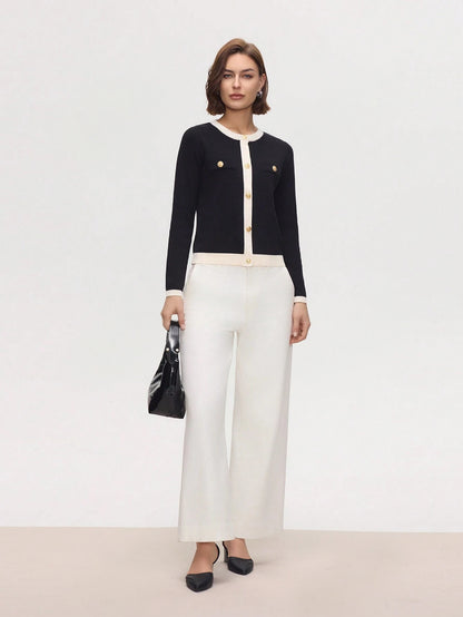 Woman wearing a black cardigan, white blouse, and white pants with a black handbag on a light gray background