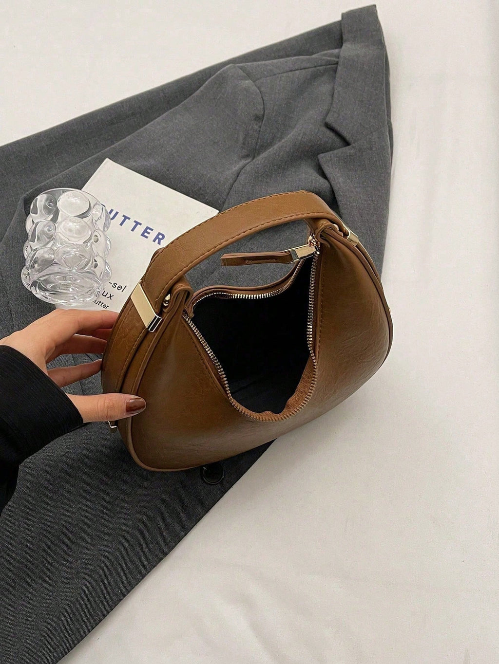 Brown leather handbag on a gray surface with a book and glass object.
