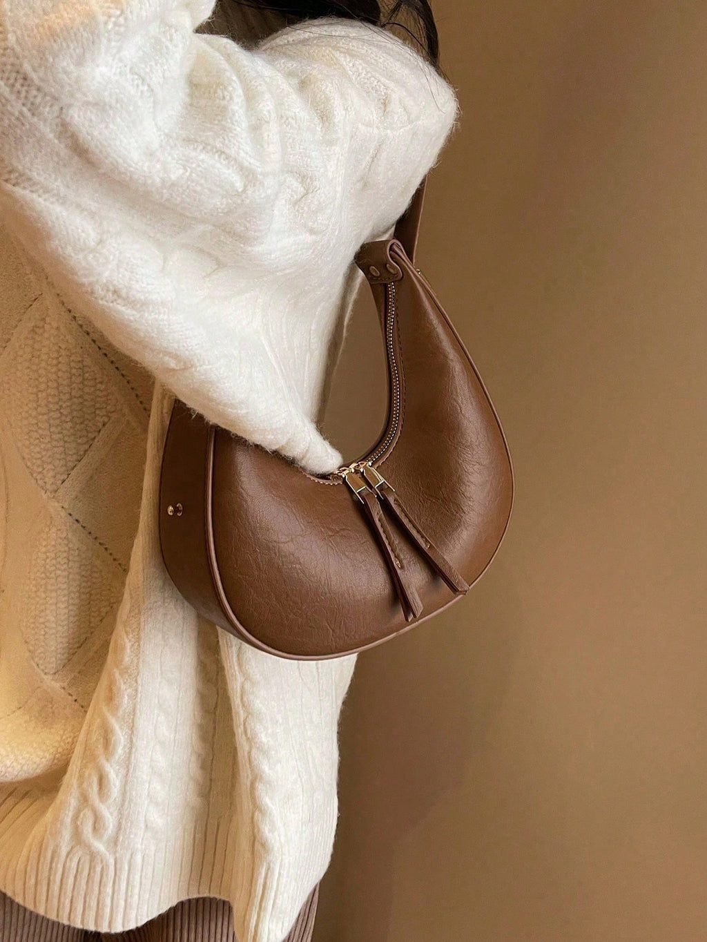 Brown leather handbag held by a person wearing a white sweater and beige coat against a beige background