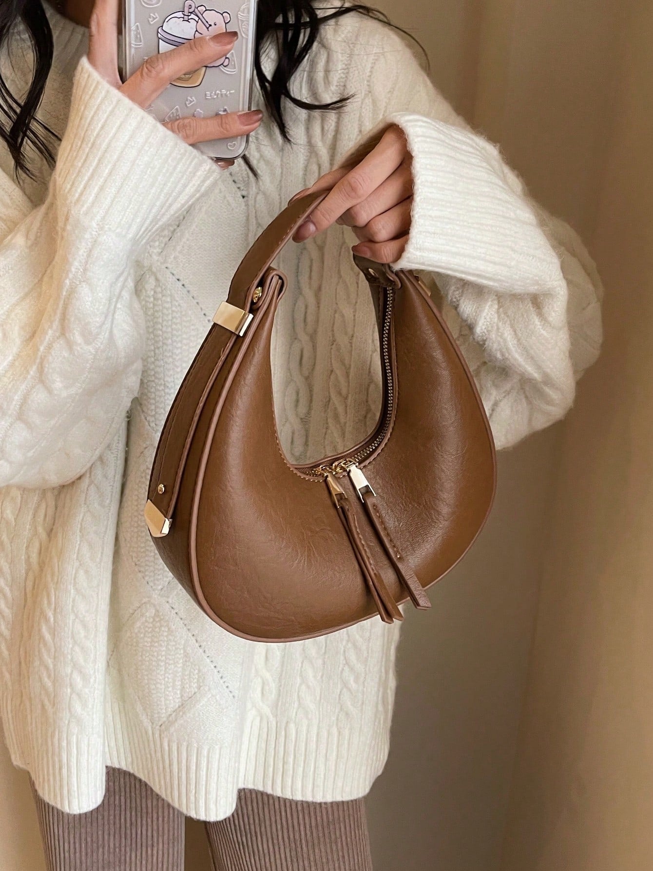 Brown handbag held by a person wearing a white sweater against a neutral background