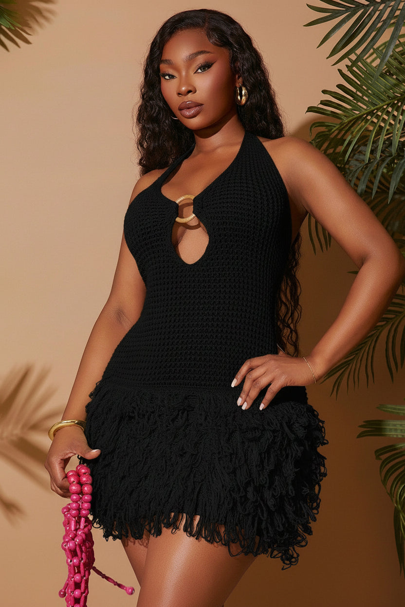 Woman wearing a black crochet dress with ruffled hem against a beige background with green foliage.