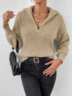 Woman wearing a beige ribbed sweater and black jeans, holding a black handbag against a white background.