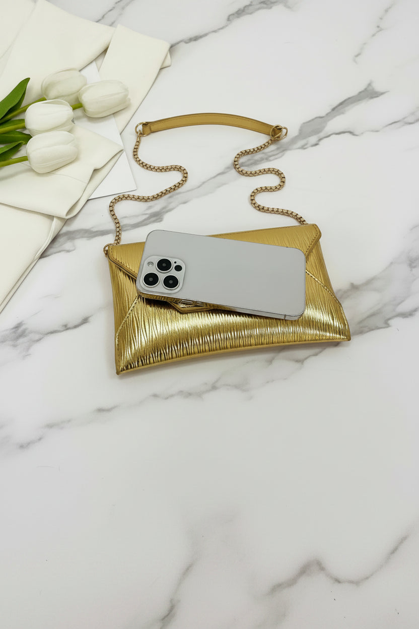 Gold clutch with chain strap holding a phone on a white surface with white tulips.
