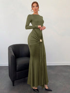 Woman wearing an olive green dress standing next to a black chair against a white wall.
