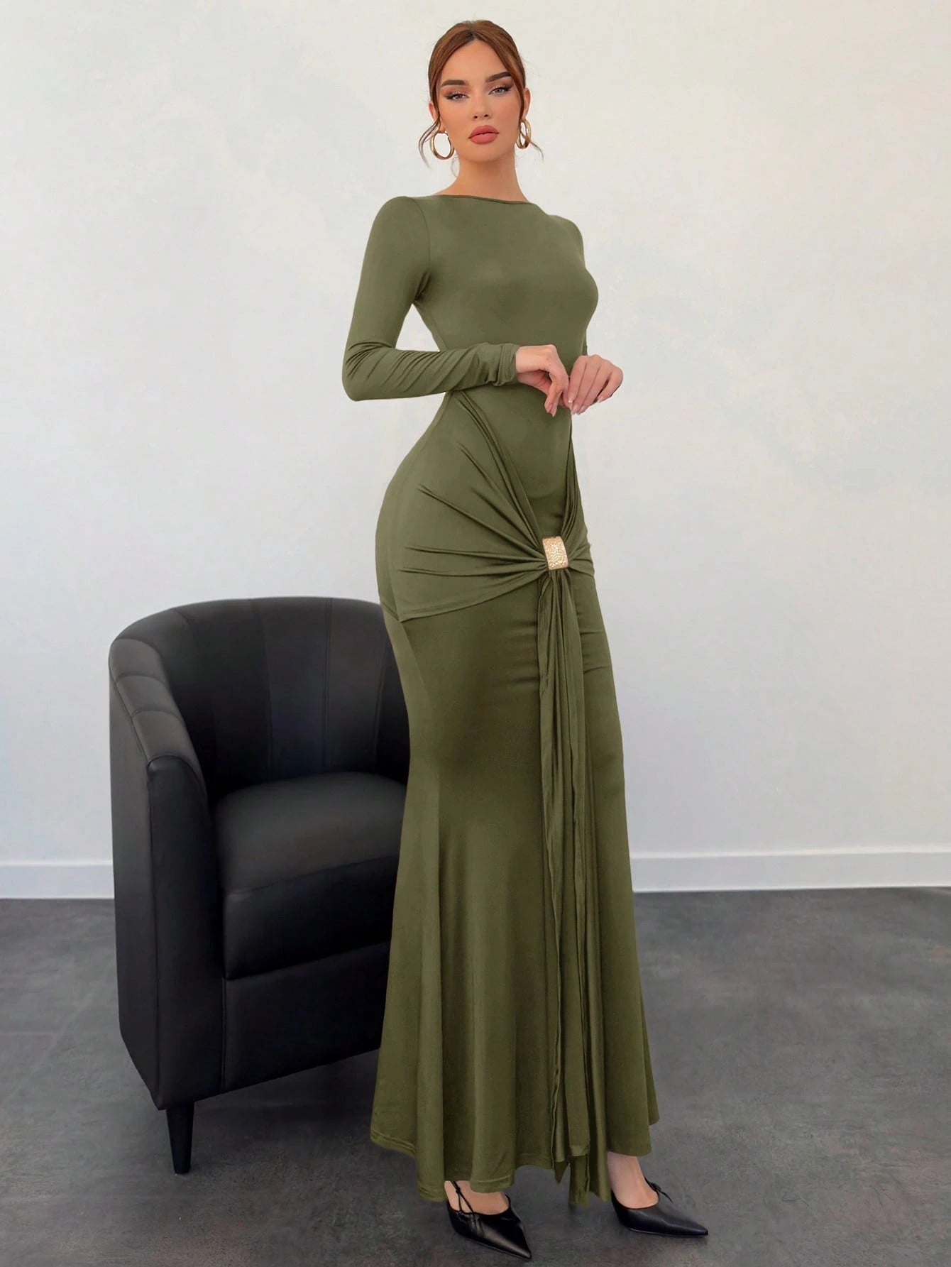 Woman wearing an olive green dress standing next to a black chair against a white wall.
