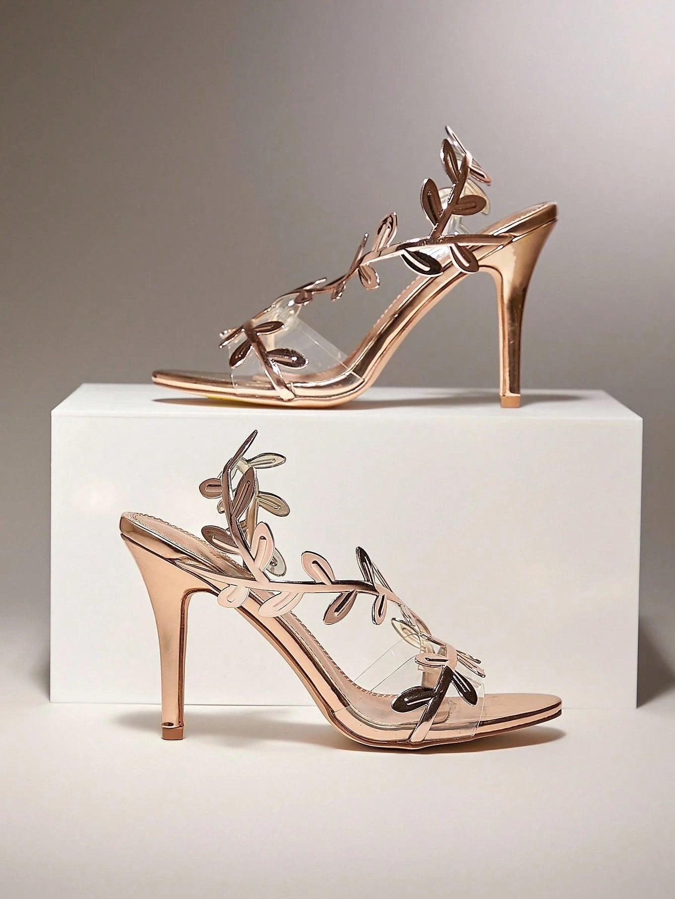 Rose gold high-heeled sandals with floral designs on a white platform against a gray background