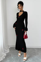 Woman wearing a black dress with floral embroidery, holding a red handbag against a white background.