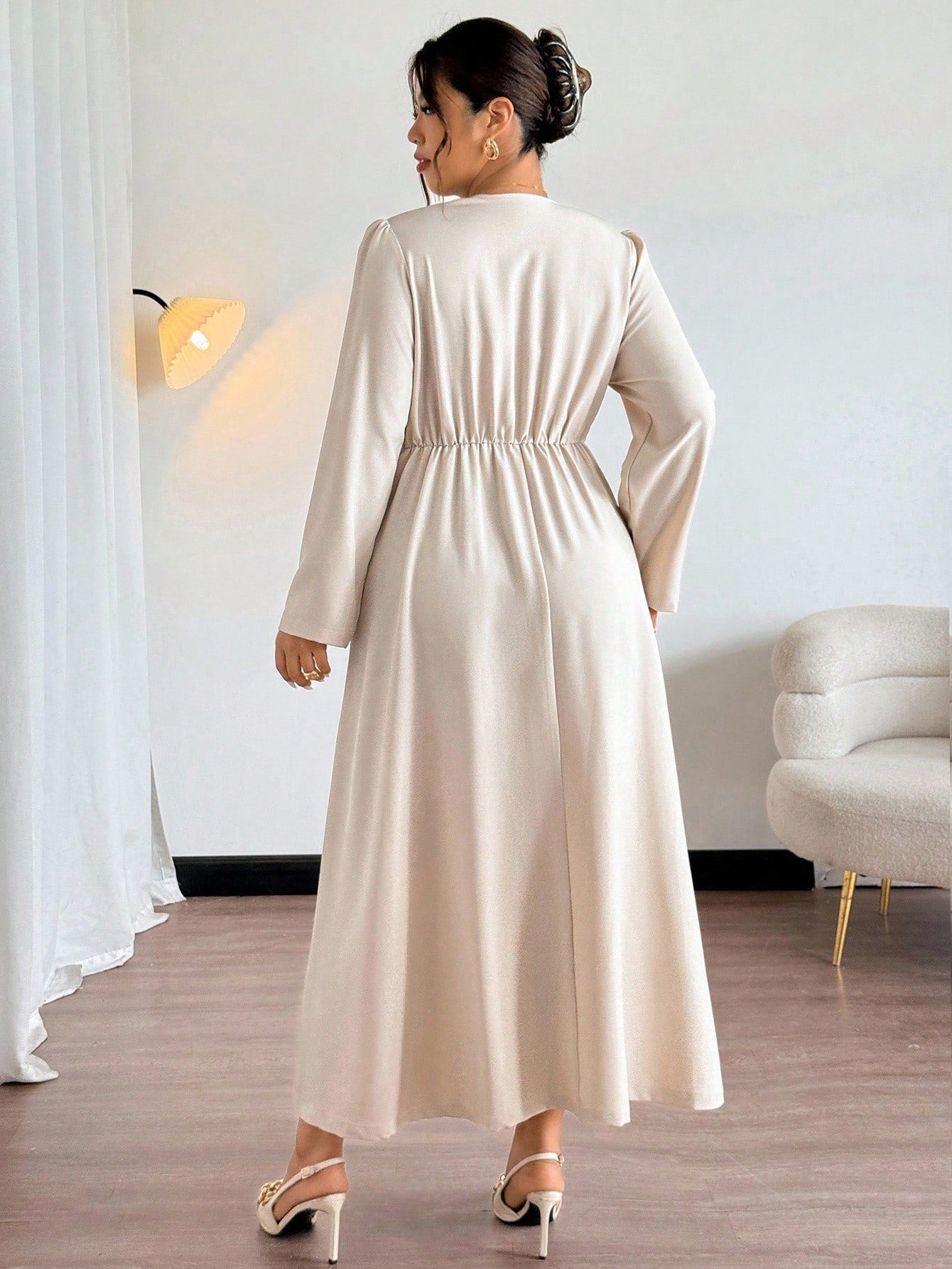 Woman wearing a long beige dress in a room with white walls and a light-colored sofa.