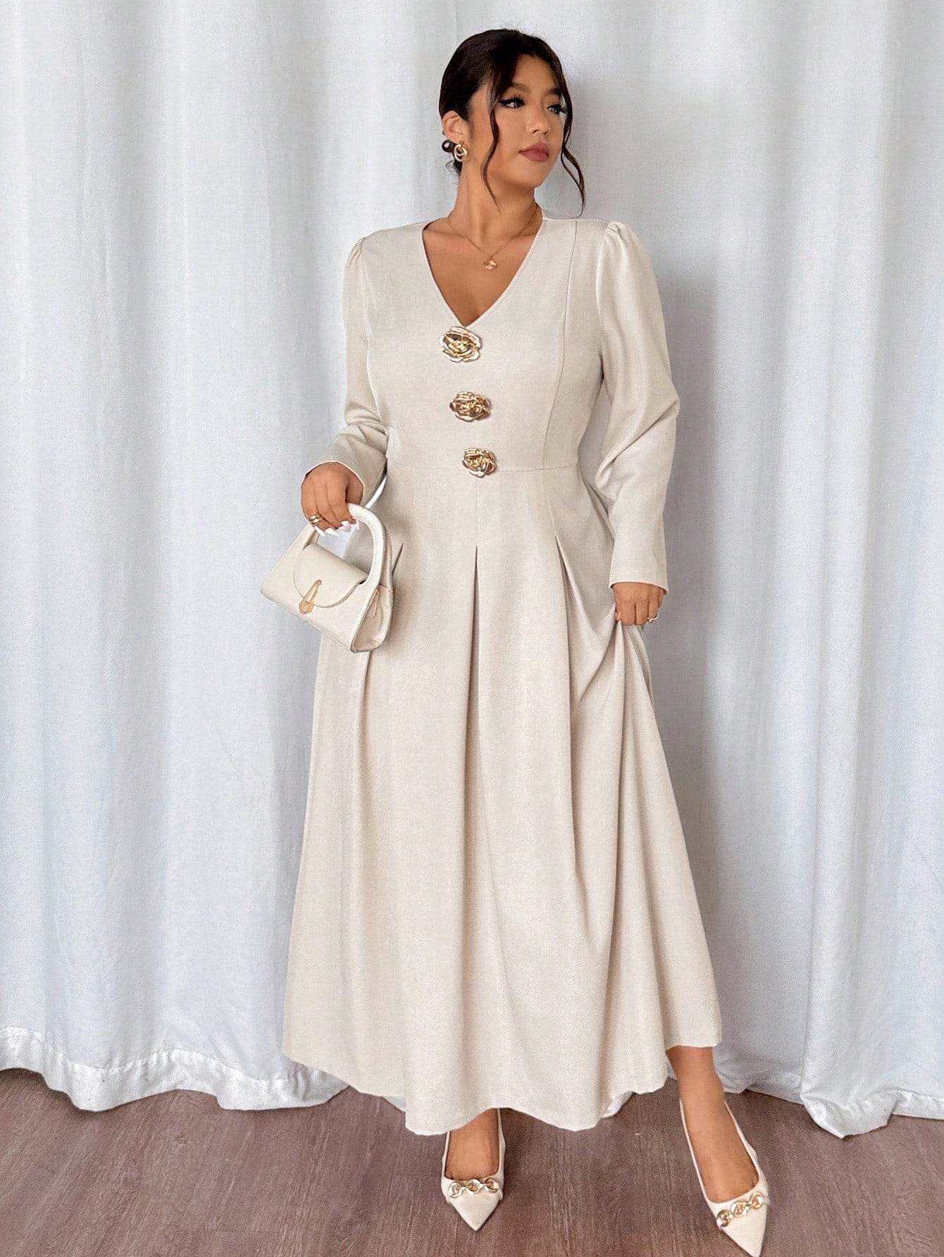 Woman wearing a long beige dress with gold buttons against a white curtain background