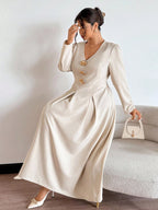 Woman in a long beige dress with gold buttons holding a white handbag against a neutral background.