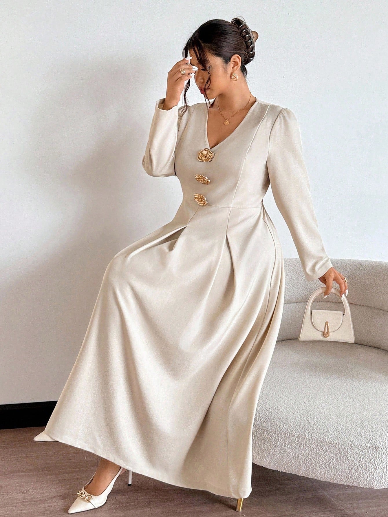 Woman in a long beige dress with gold buttons holding a white handbag against a neutral background.