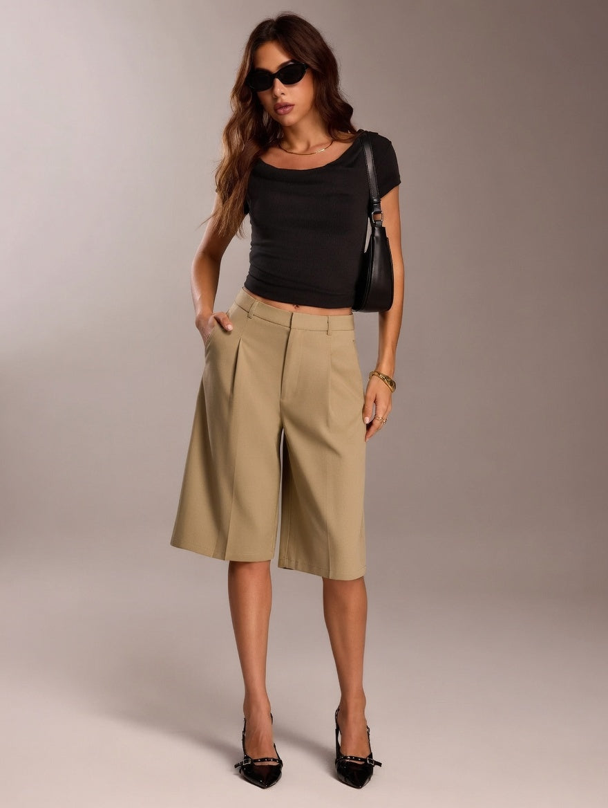 Woman wearing a black top and beige shorts with a black handbag against a plain background