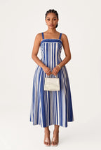 Woman wearing an A-line midi dress features a bold, vertical stripe pattern in alternating royal blue and white that creates a slimming and elongating visual effect. Designed with a structured square neckline, thick supportive straps, and a solid blue upper border, the fitted bodice gracefully transitions into a flared, flowing skirt that falls just below the calves.